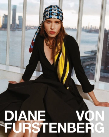 Model wearing a black dress and patterned headscarf sitting indoors against a large window overlooking a city skyline at dusk.