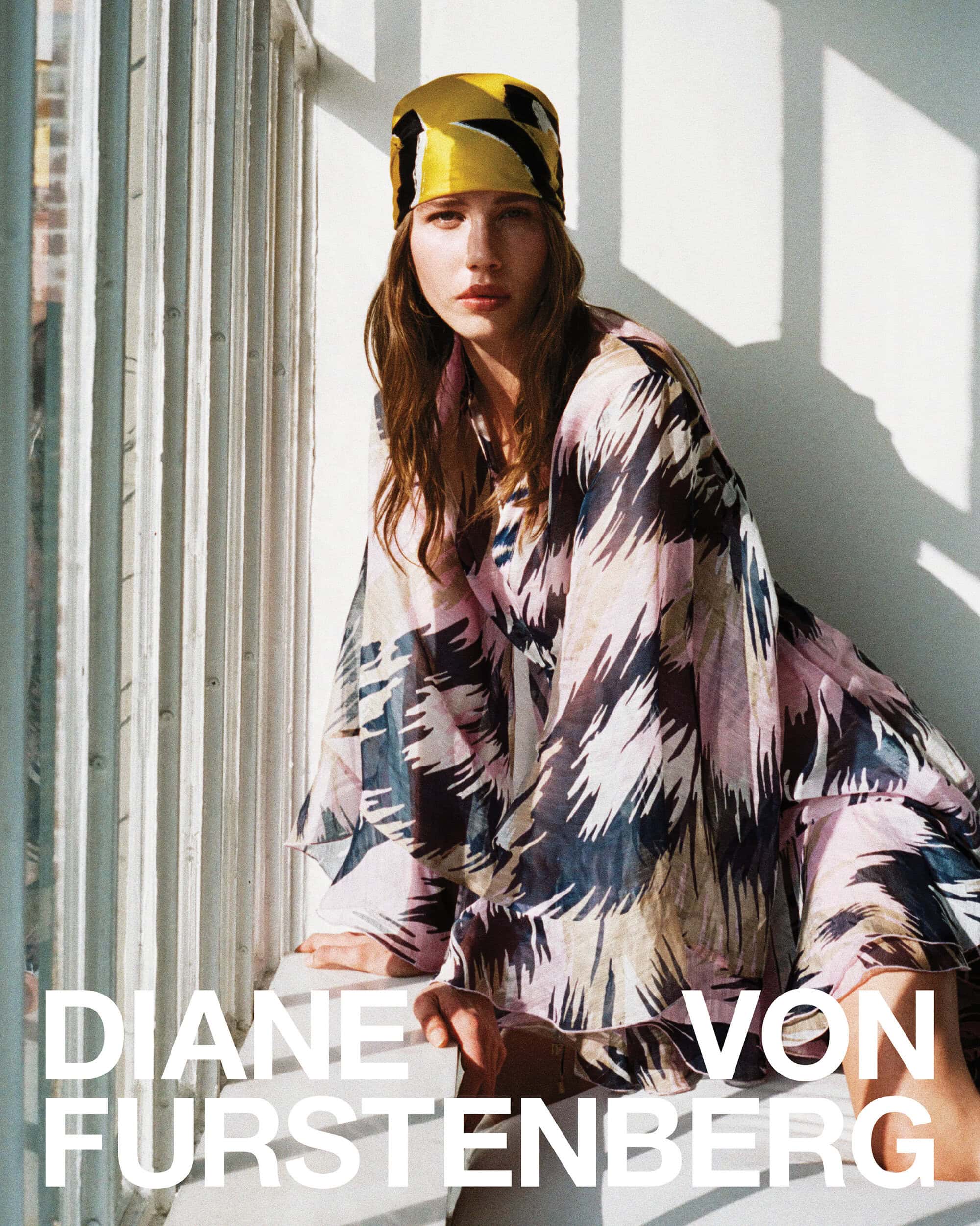 Model wearing a patterned silk headscarf and matching dress posed near a window with harsh morning sunlight casting shadows.