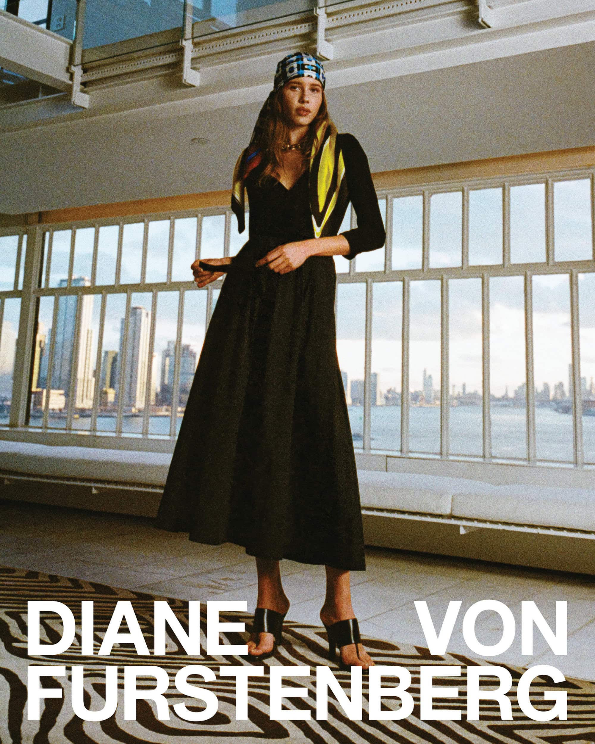 Model wearing black dress and headscarf standing in interior space with large windows overlooking a city skyline and water.