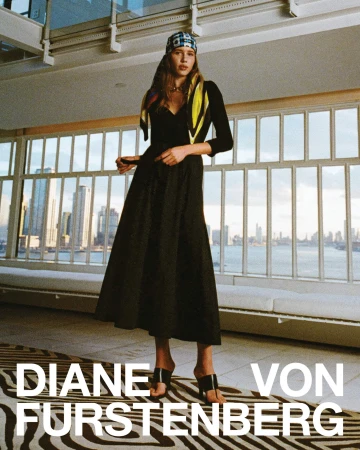 Model wearing black dress and headscarf standing in interior space with large windows overlooking a city skyline and water.