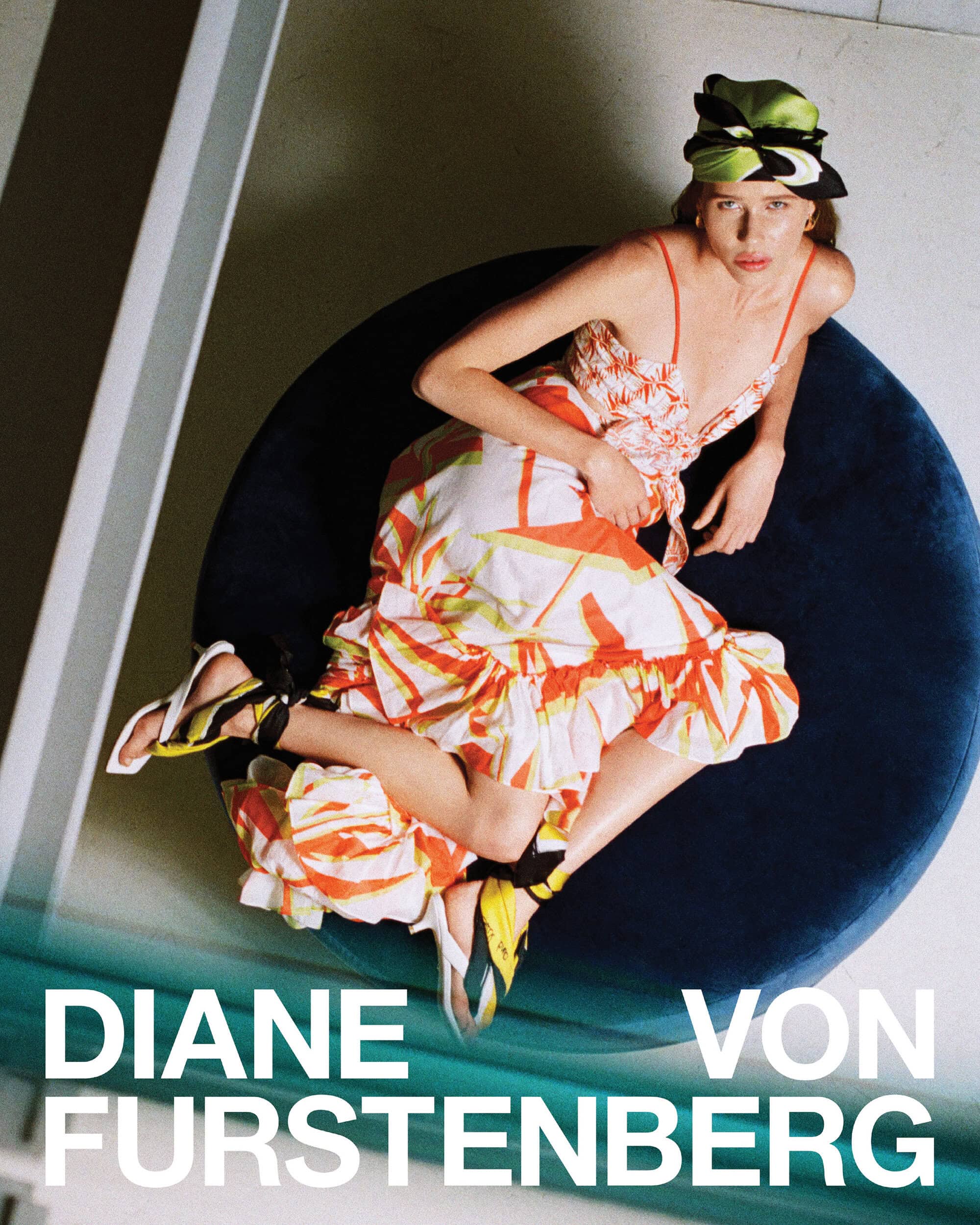 Model in printed dress and patterned headscarf reclining on a dark blue circular lounge chair for Diane von Furstenberg