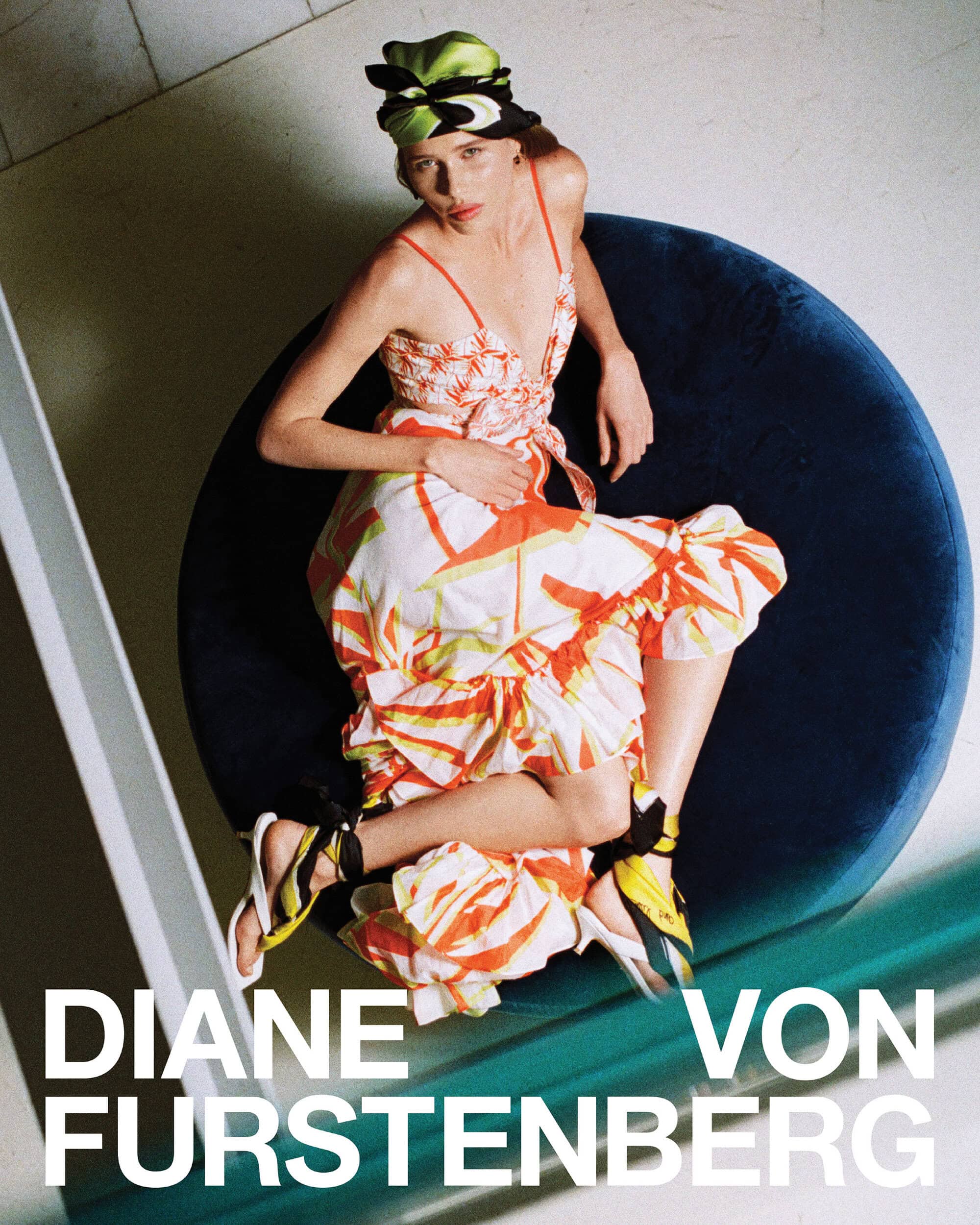 Model seated on a round blue velvet cushion wearing a patterned orange and white dress and head wrap