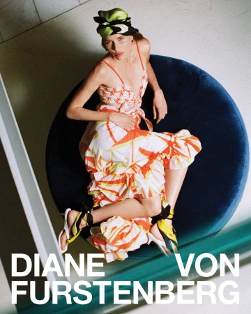 Model seated on a round blue velvet cushion wearing a patterned orange and white dress and head wrap
