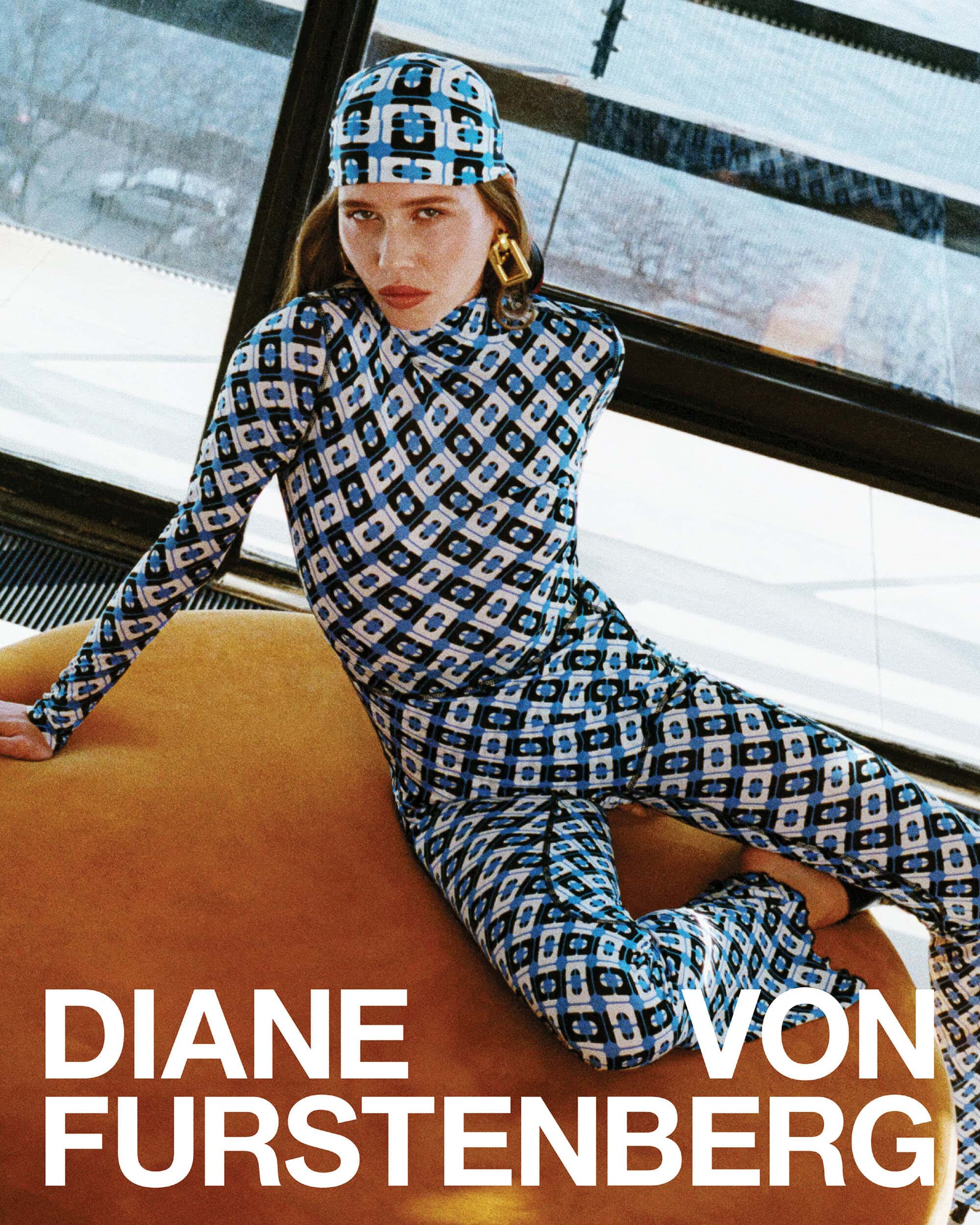 Model in blue patterned turtleneck and trousers with matching headscarf seated on an orange round ottoman