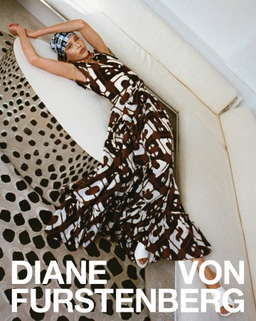 Model in printed wrap dress and headscarf reclining against a curved white sofa over patterned carpet.