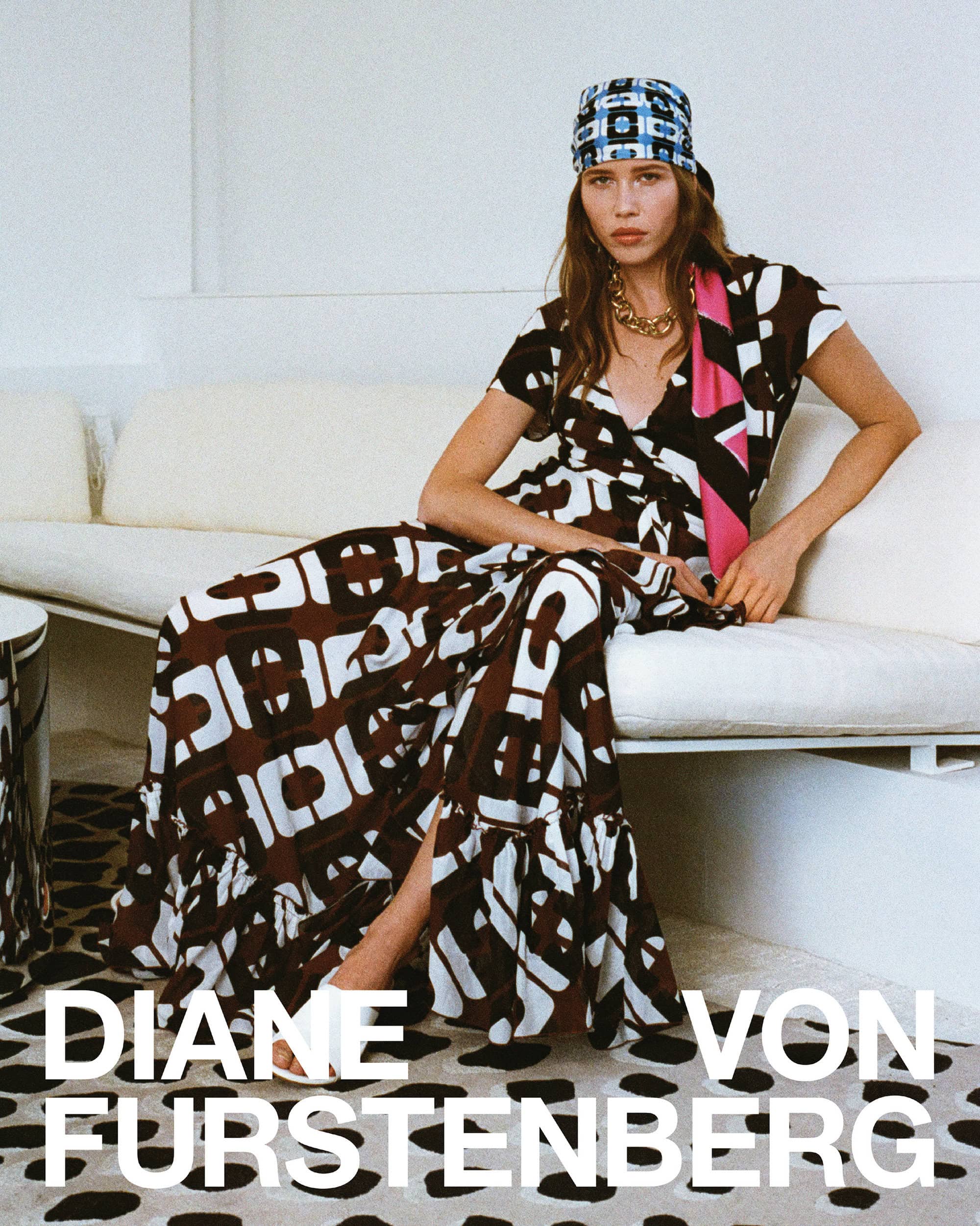 Model seated on a white sofa wearing a patterned wrap dress and printed headscarf for Diane von Furstenberg