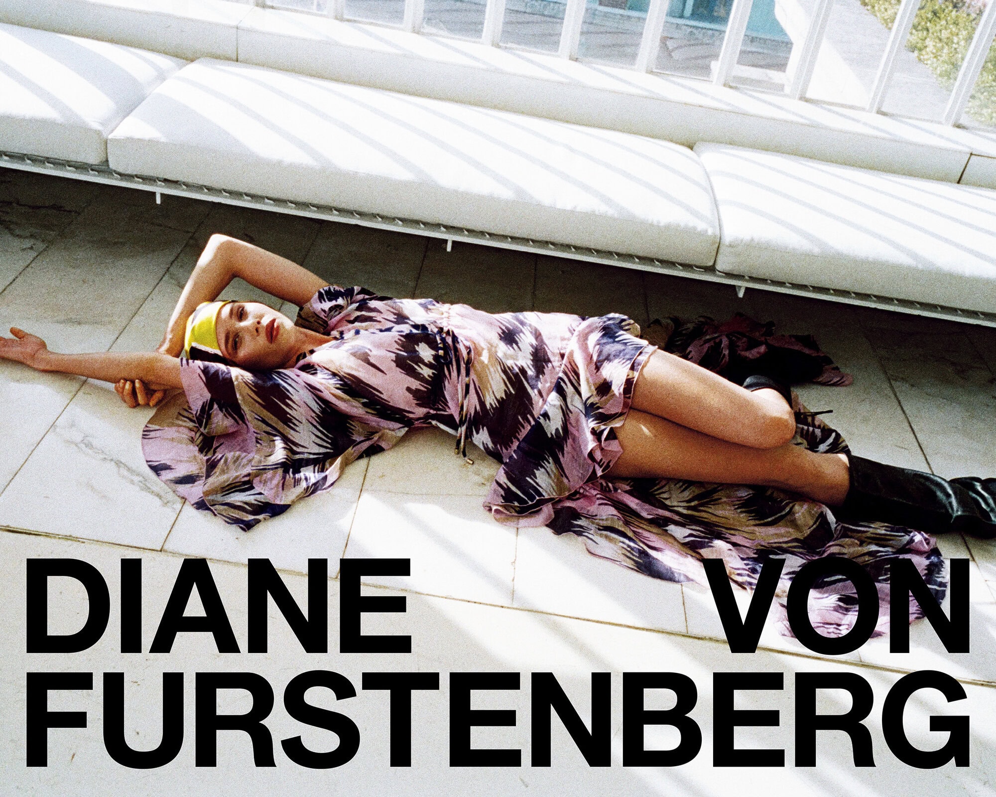 Model lying on tiled floor wearing a patterned dress below a white sofa for Diane von Furstenberg