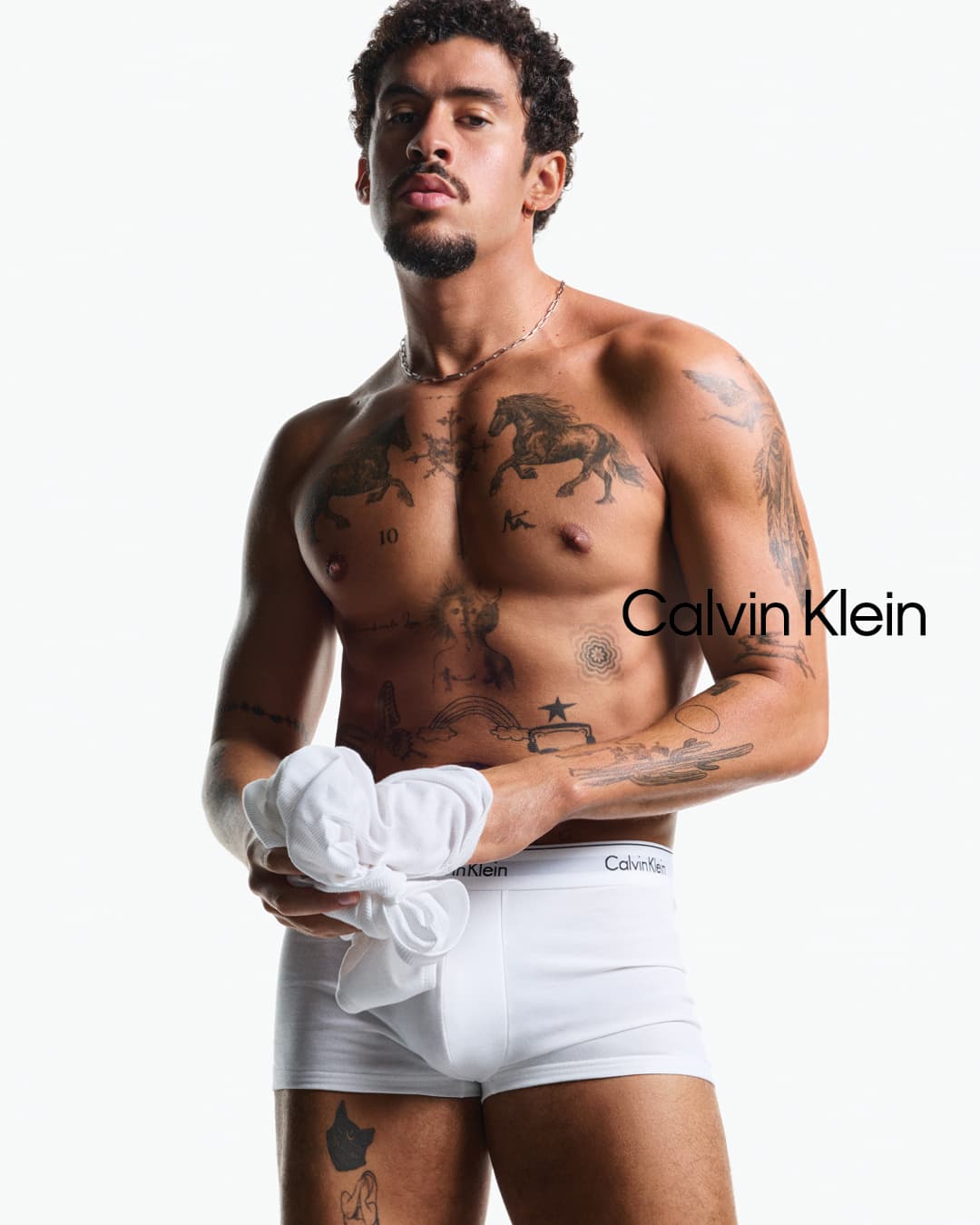 Bad Bunny shirtless wearing white underwear holding a shirt against a seamless white background for Calvin Klein