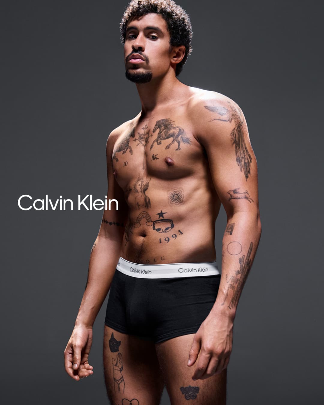 Bad Bunny posing shirtless in black Calvin Klein boxer briefs against a dark grey studio background.