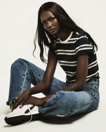 Black model seated wearing striped t-shirt and blue jeans against white background