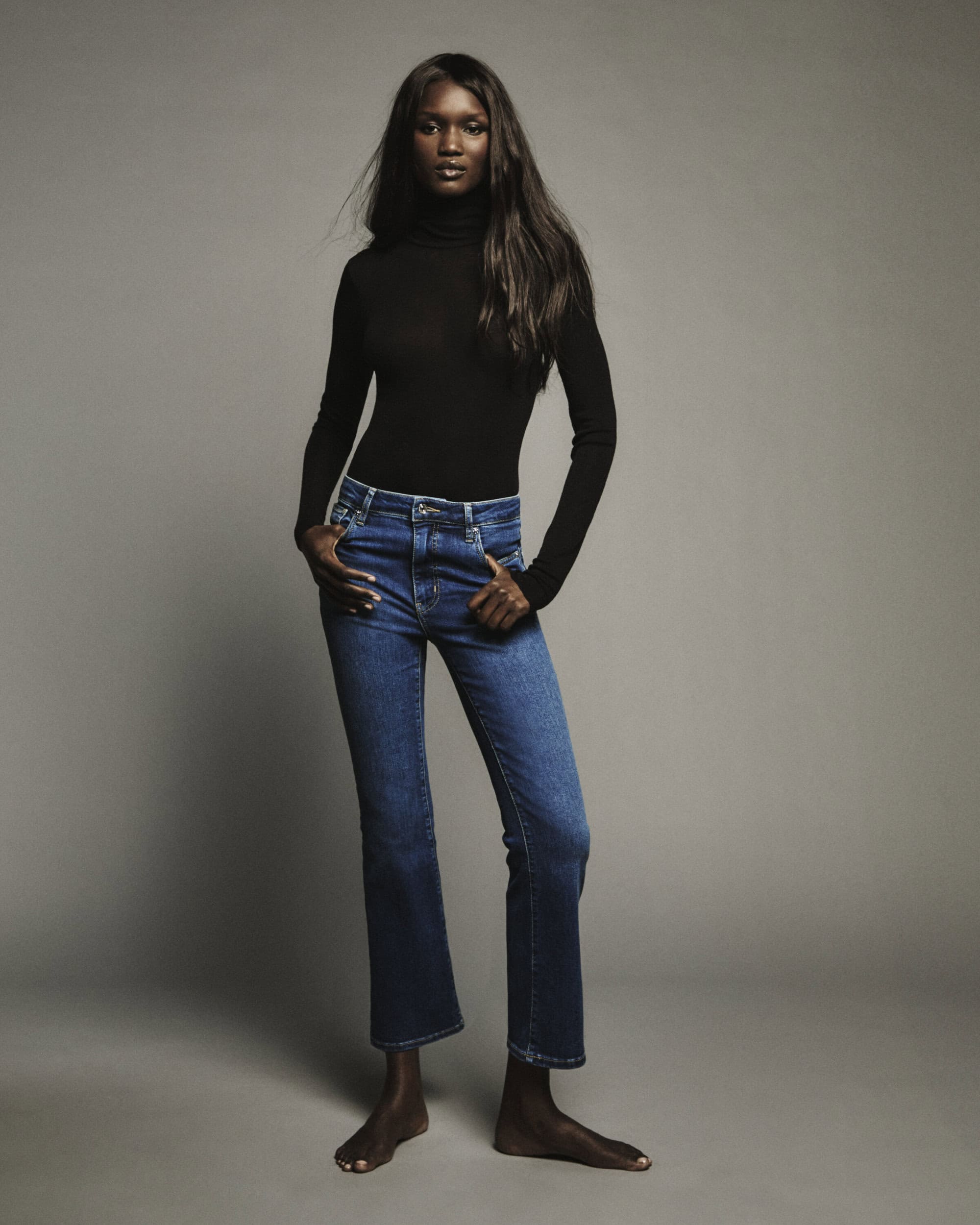 Model standing against a plain grey background wearing a black turtleneck and blue denim jeans.