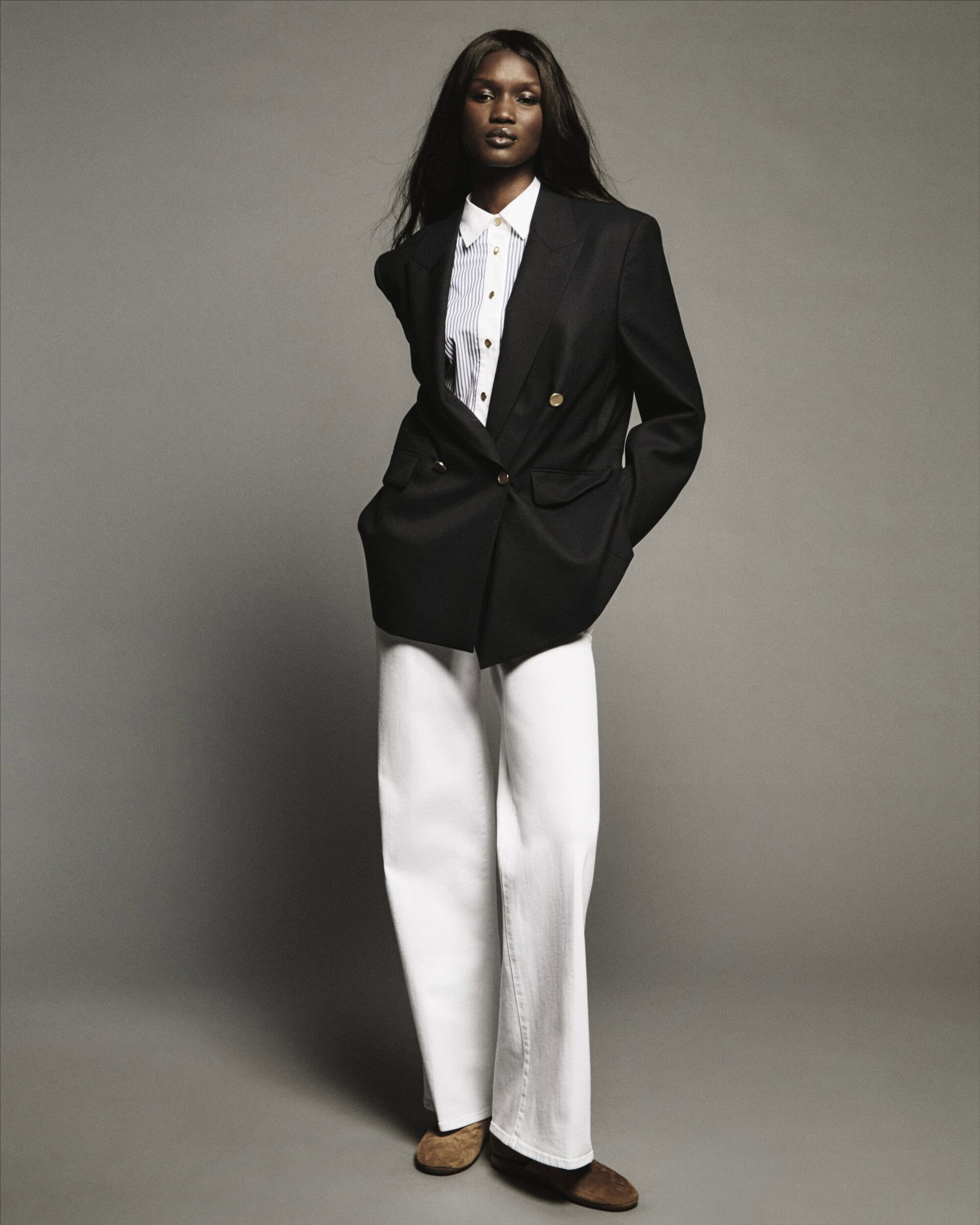 Model standing against a grey backdrop wearing a black blazer, white shirt, and white trousers.