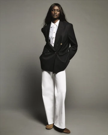 Model standing against a grey backdrop wearing a black blazer, white shirt, and white trousers.