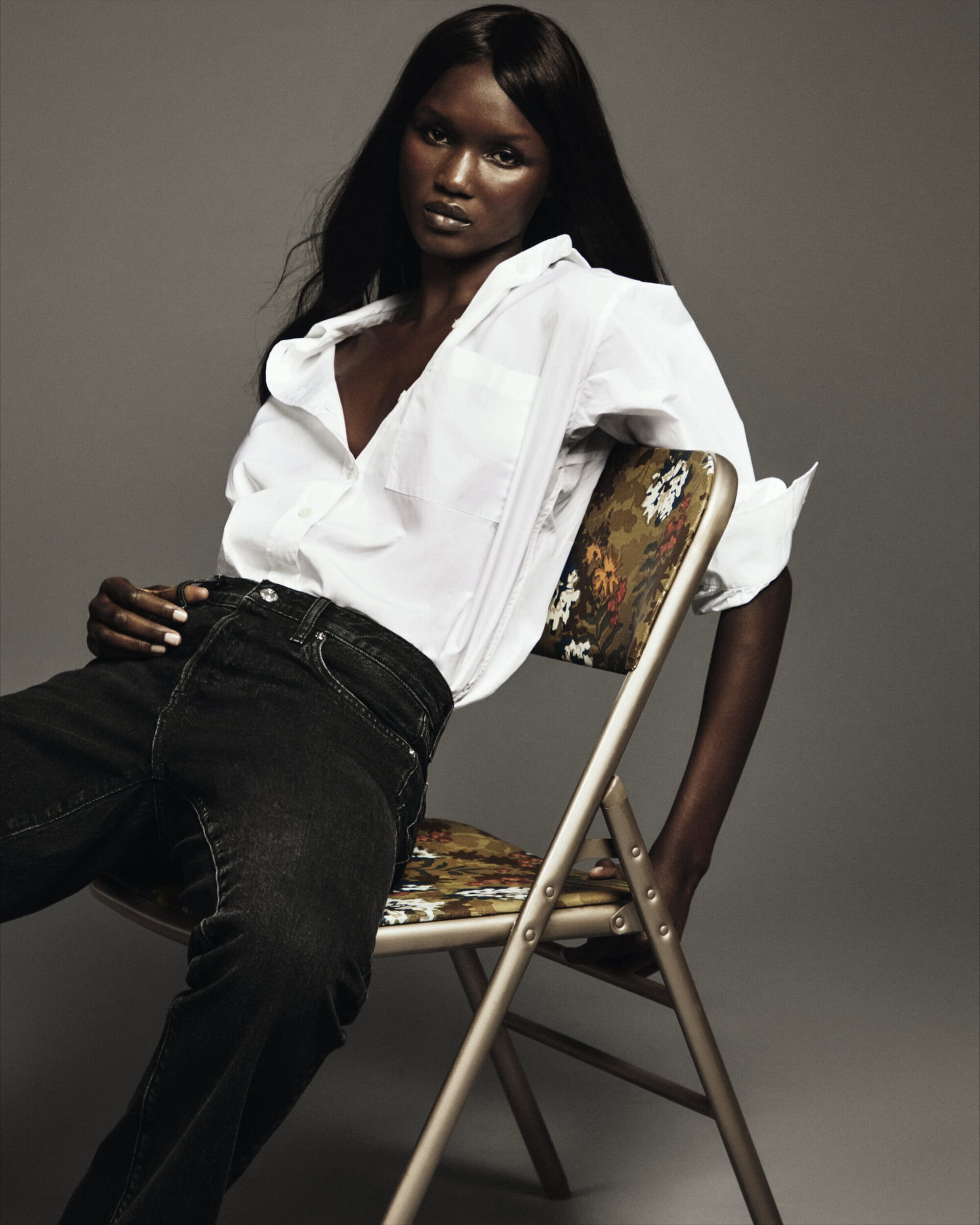 Model with long dark hair seated on a folding chair wearing a white shirt and black jeans against a grey background
