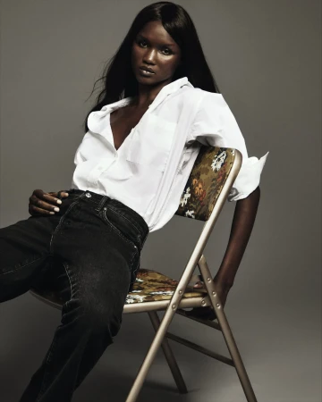 Model with long dark hair seated on a folding chair wearing a white shirt and black jeans against a grey background