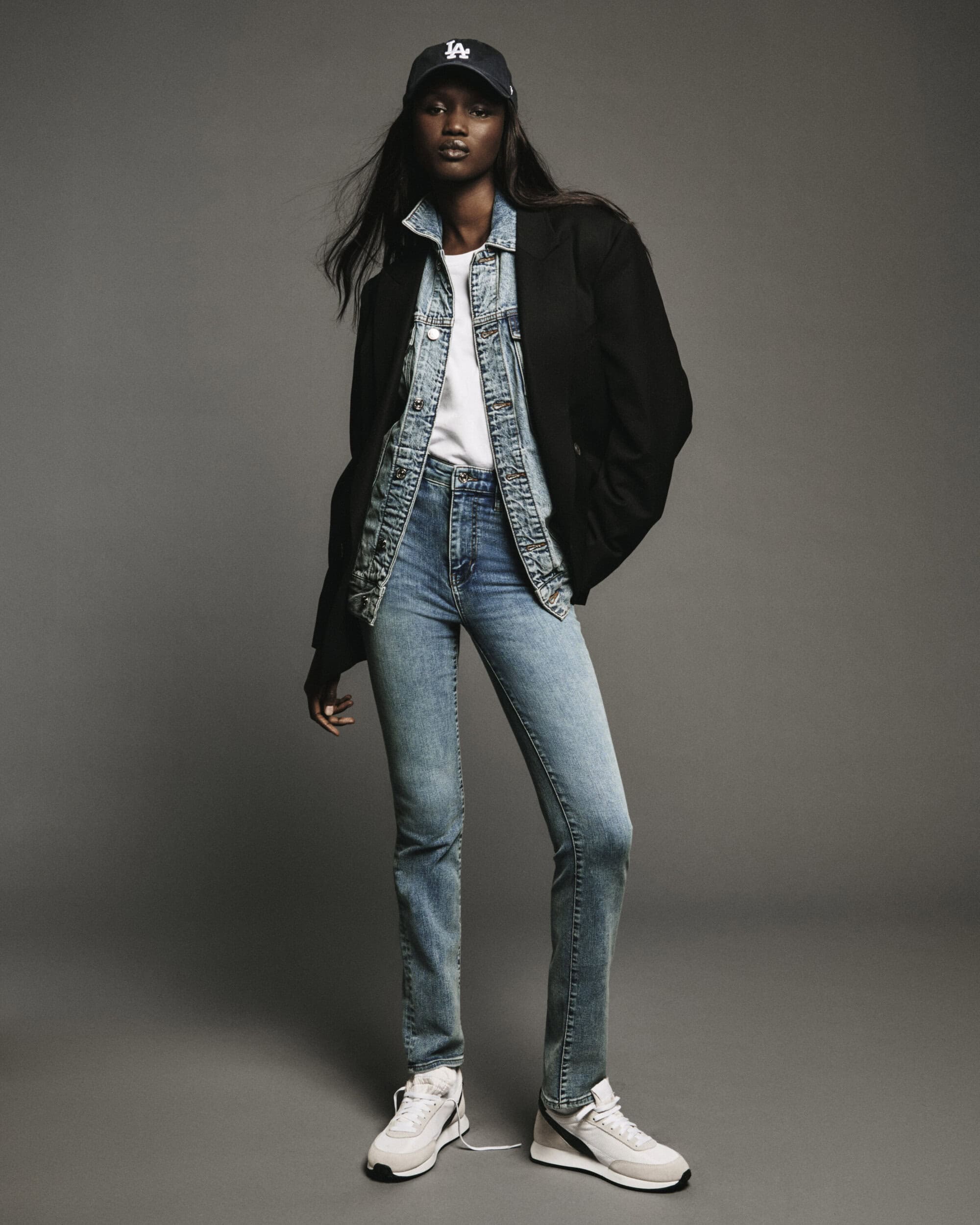 Model wearing blue jeans, denim jacket, black blazer, white t-shirt, and baseball cap standing against grey seamless backdrop