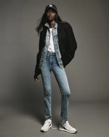 Model wearing blue jeans, denim jacket, black blazer, white t-shirt, and baseball cap standing against grey seamless backdrop