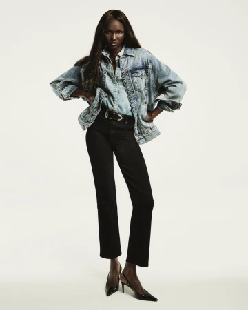 Model wearing a denim jacket and black jeans with hands on hips against a plain white studio background.