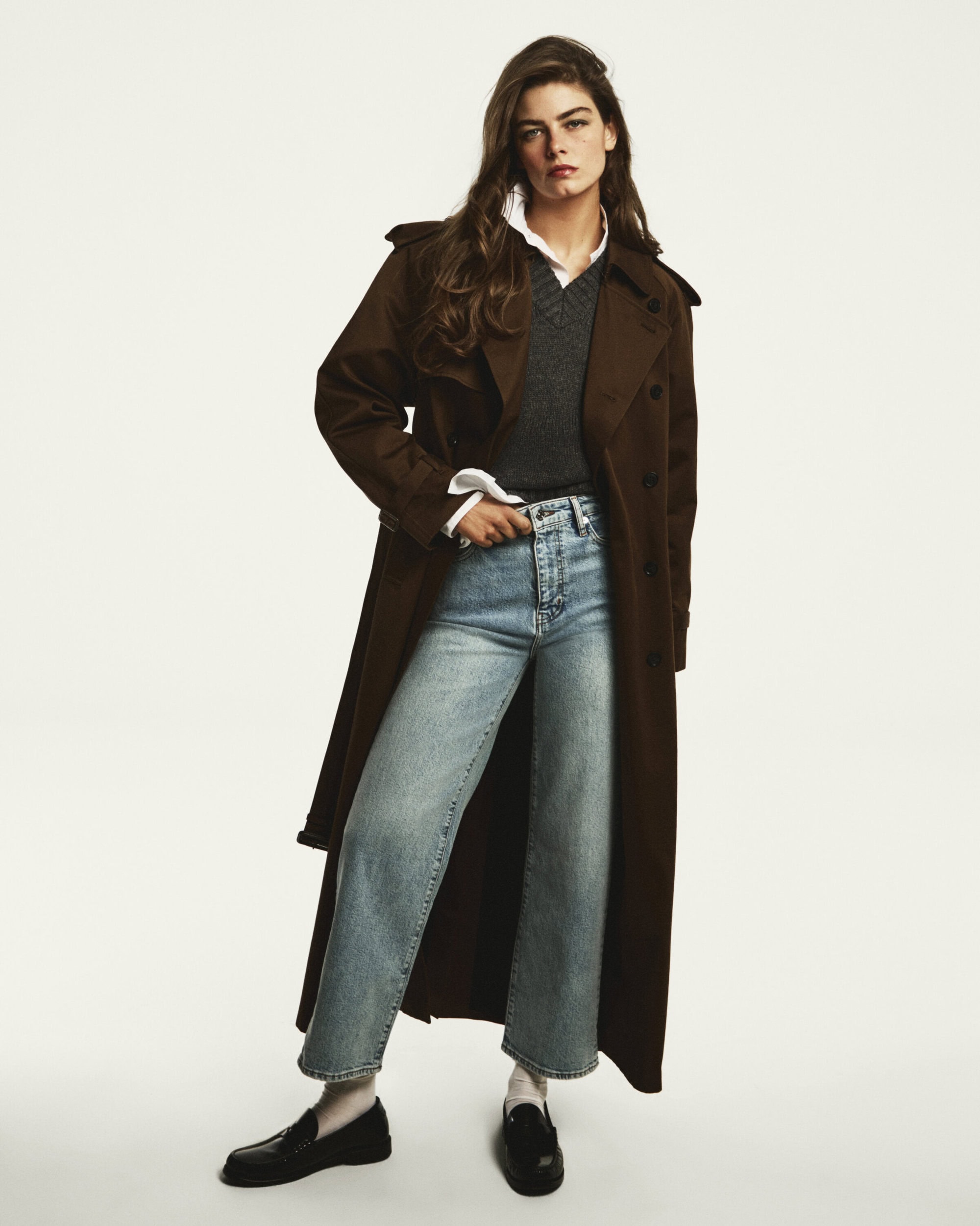Model wearing a long brown trench coat, grey v-neck sweater, and light wash denim jeans against a seamless white background.