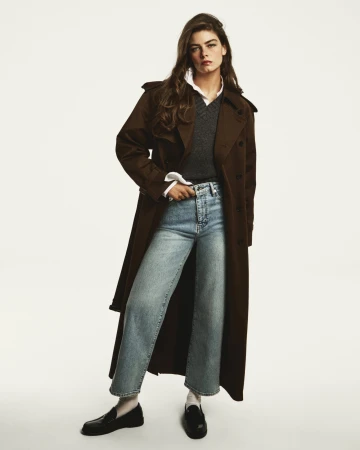 Model wearing a long brown trench coat, grey v-neck sweater, and light wash denim jeans against a seamless white background.