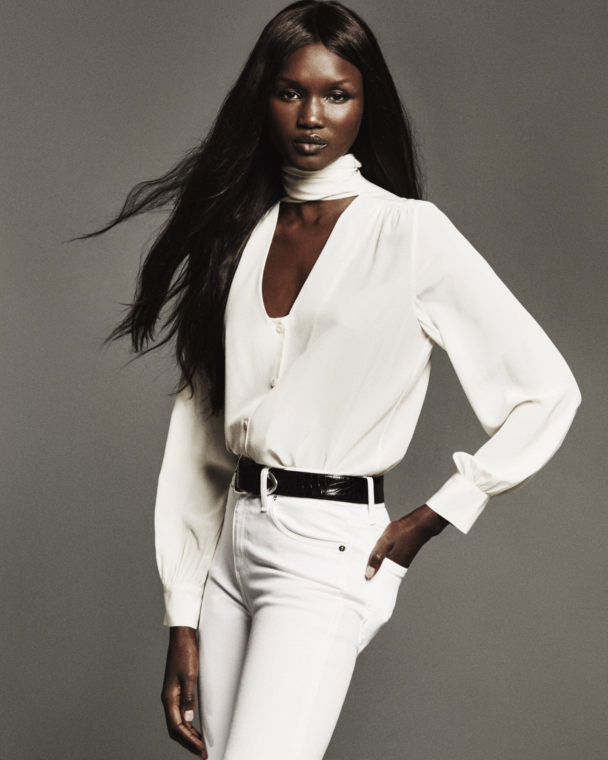 Model with long dark hair wearing a white blouse and white jeans against a grey studio backdrop