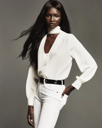 Model with long dark hair wearing a white blouse and white jeans against a grey studio backdrop