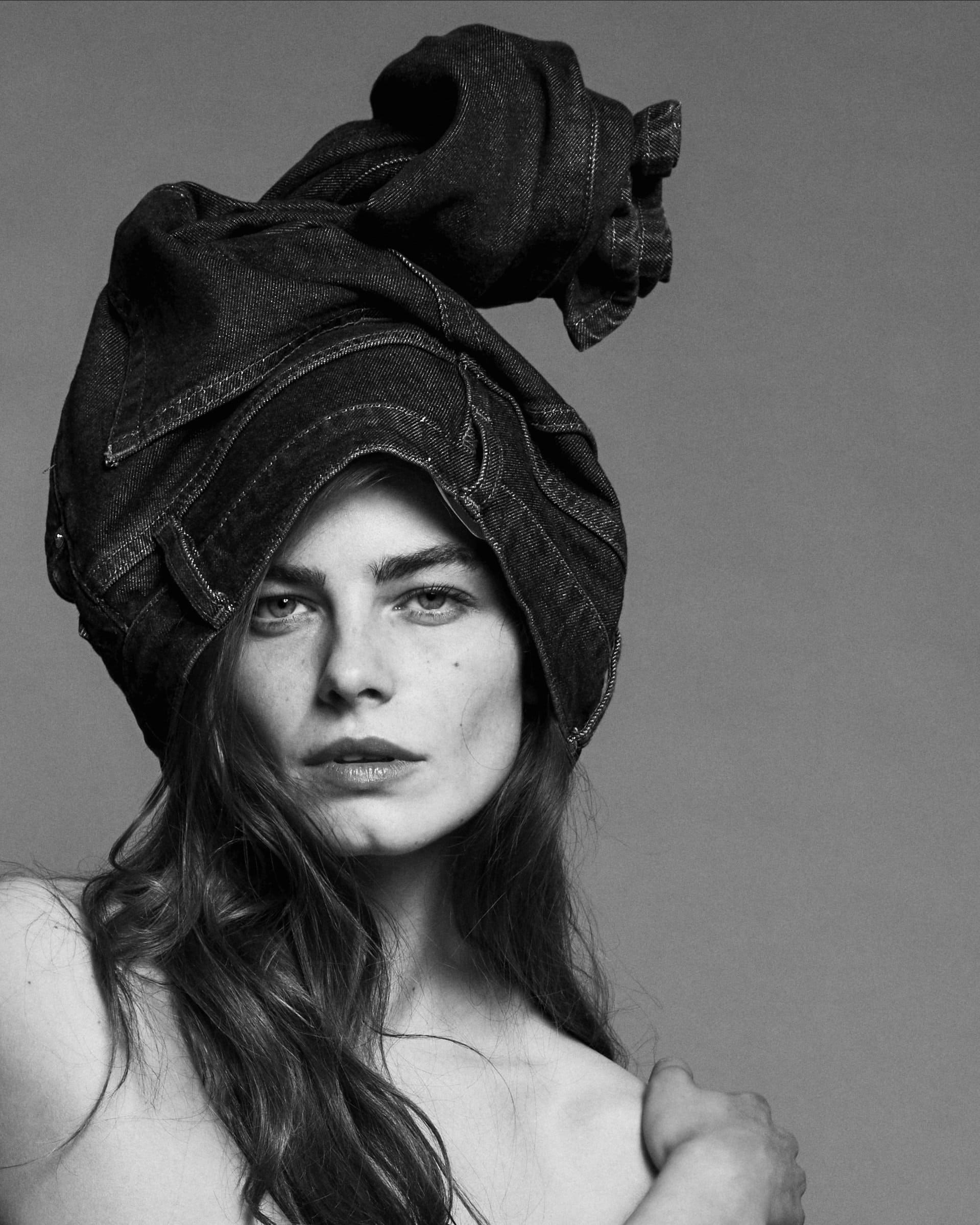 Model with long dark hair wearing dark denim jeans wrapped around her head like a hat against a grey studio background.