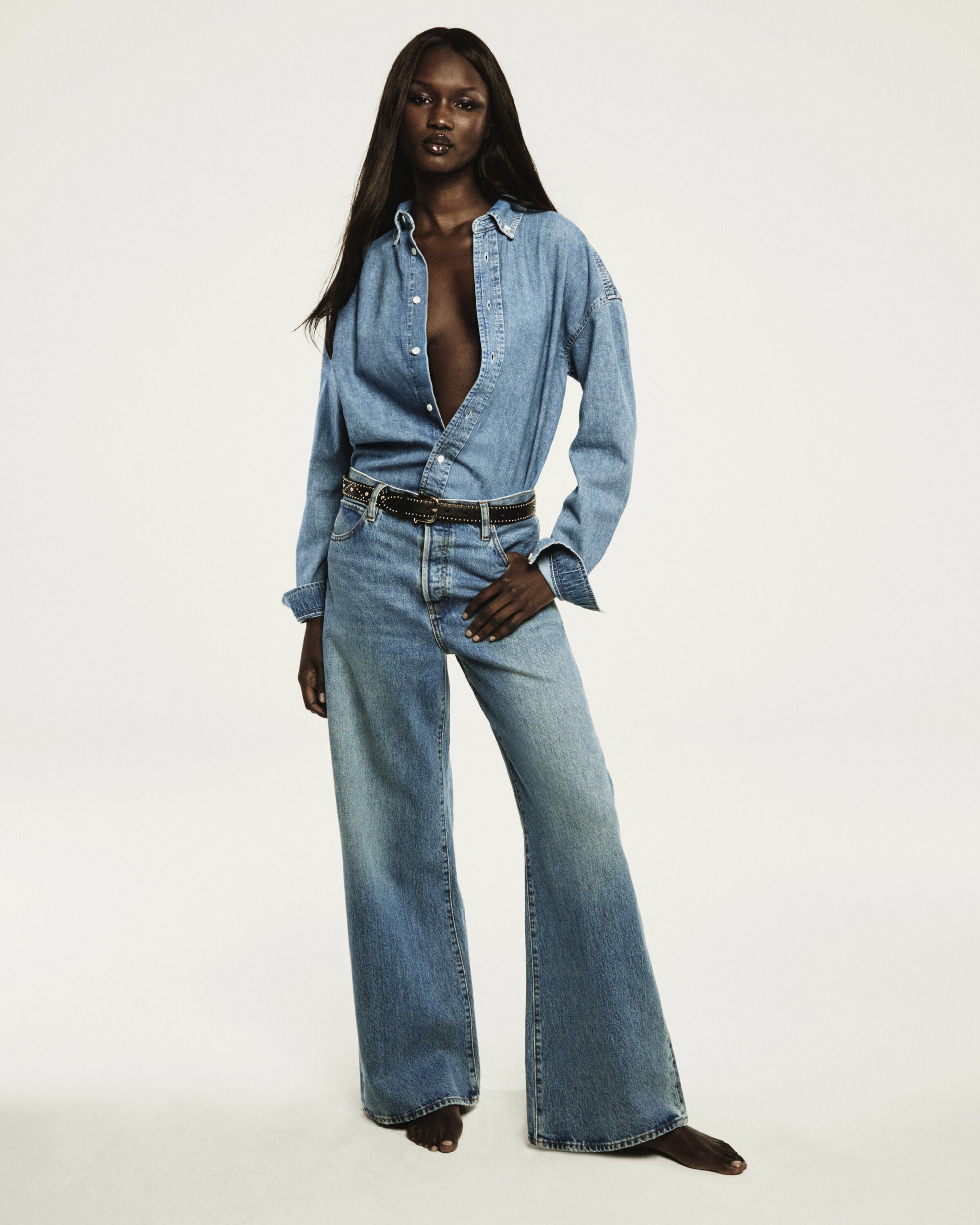 A Black woman with long dark hair wearing a denim shirt and jeans posing against a seamless white background for FRAME.