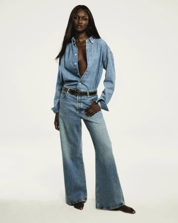 A Black woman with long dark hair wearing a denim shirt and jeans posing against a seamless white background for FRAME.