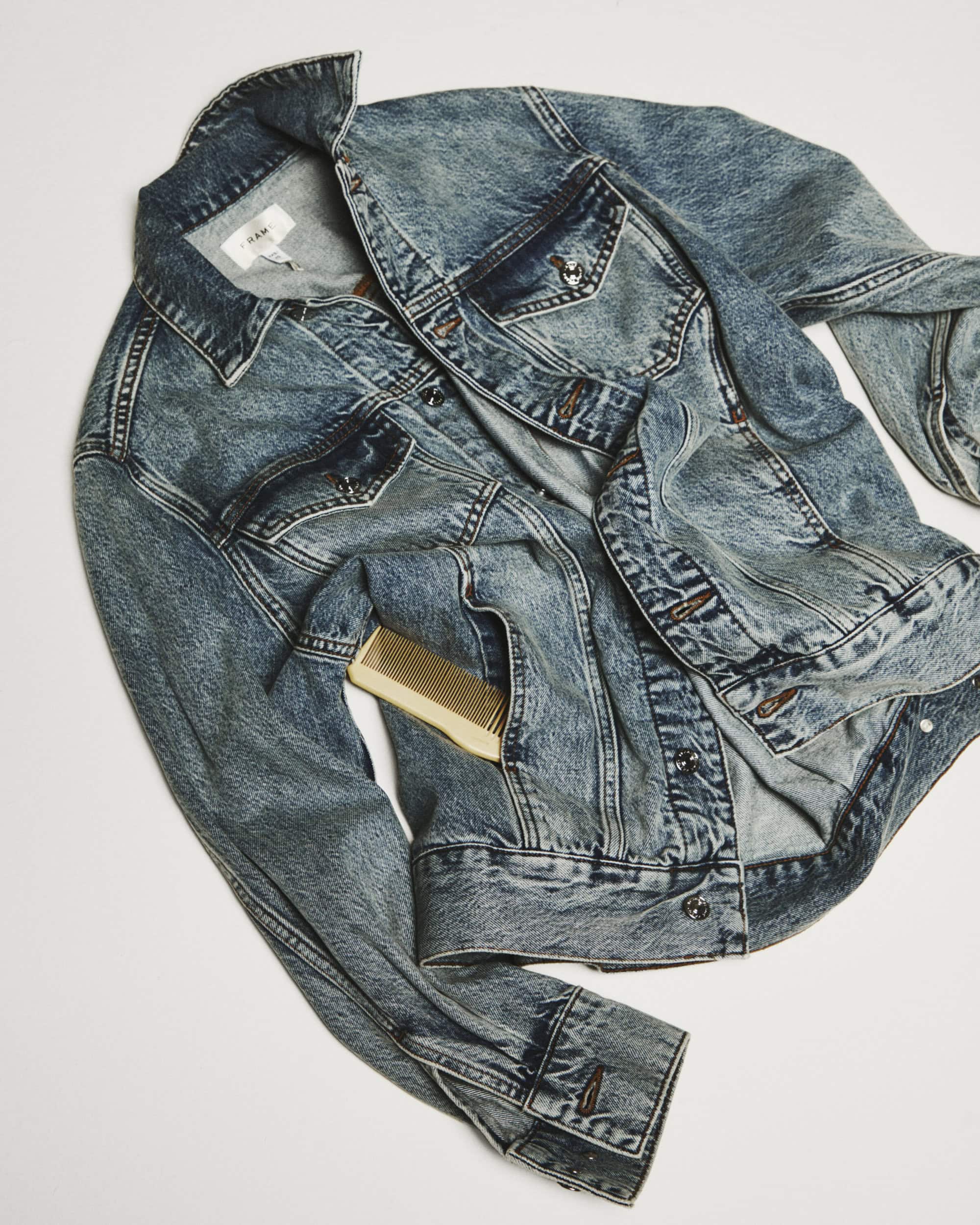 Acid wash denim jacket with a small comb tucked into the pocket against a neutral background for FRAME spring campaign