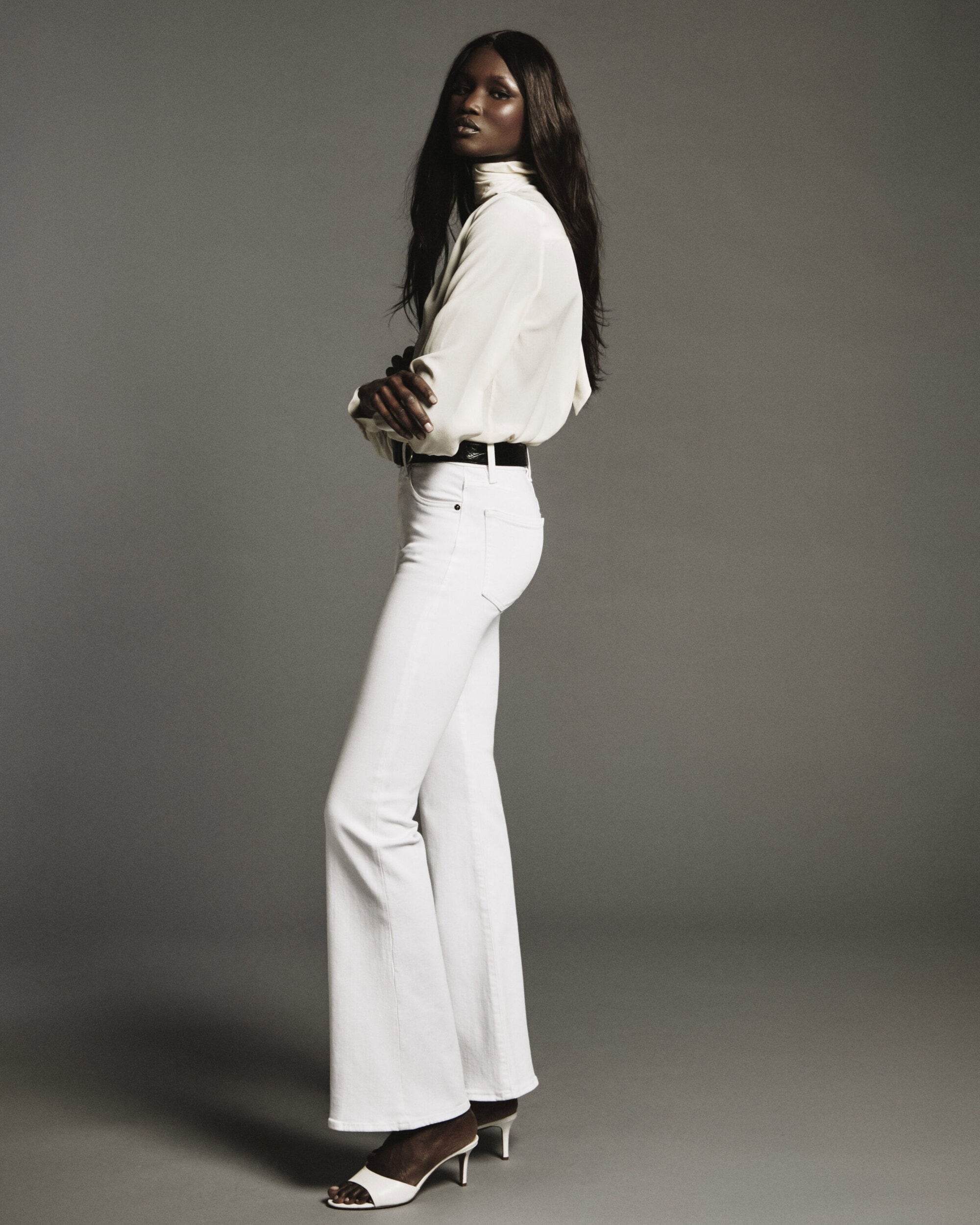 A female model in white blouse and white denim pants posing against a neutral grey seamless background.