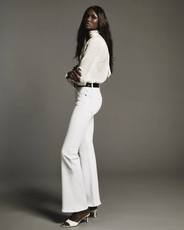 A female model in white blouse and white denim pants posing against a neutral grey seamless background.