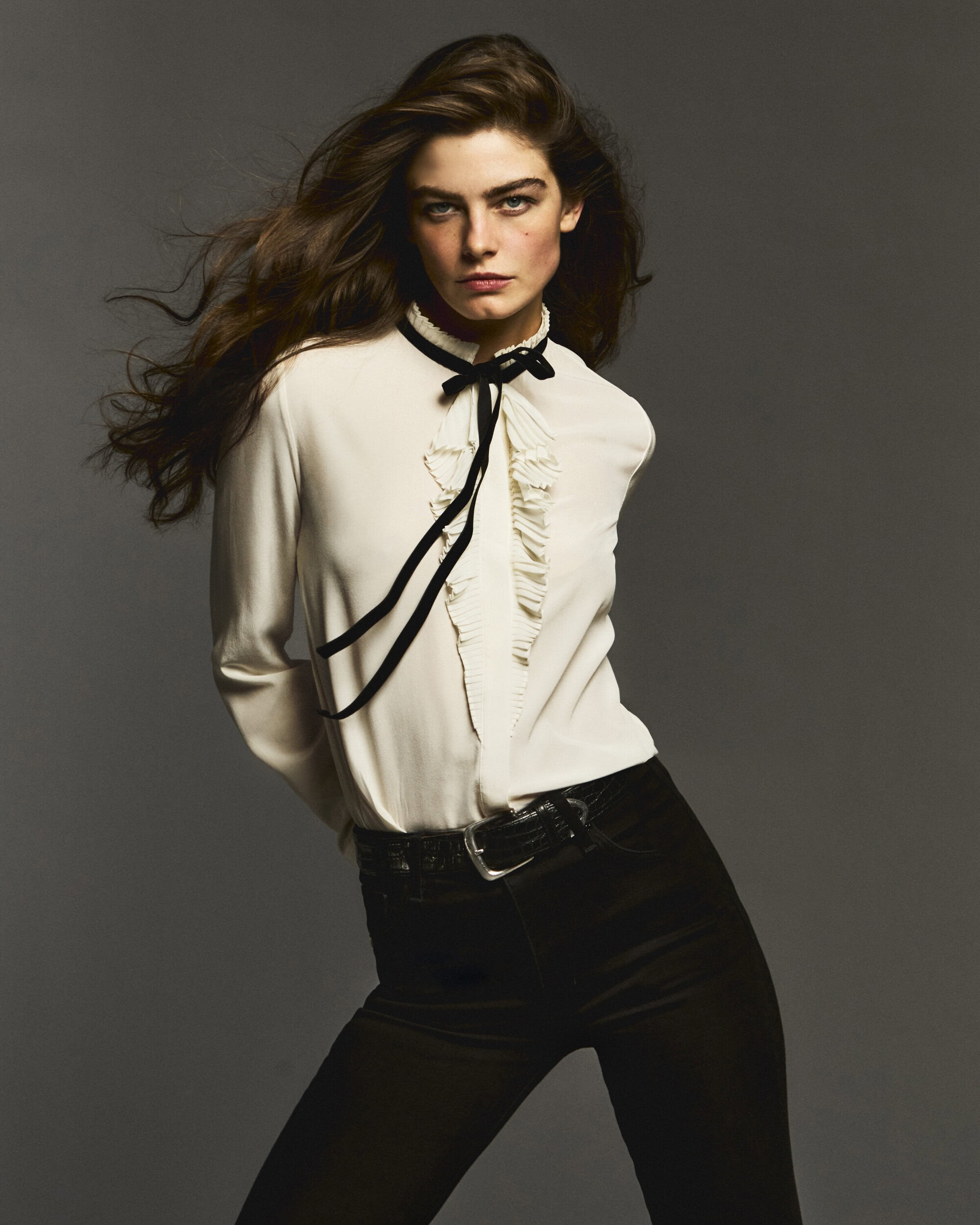 Model with long dark hair wearing a white ruffled blouse and black trousers against a neutral grey background