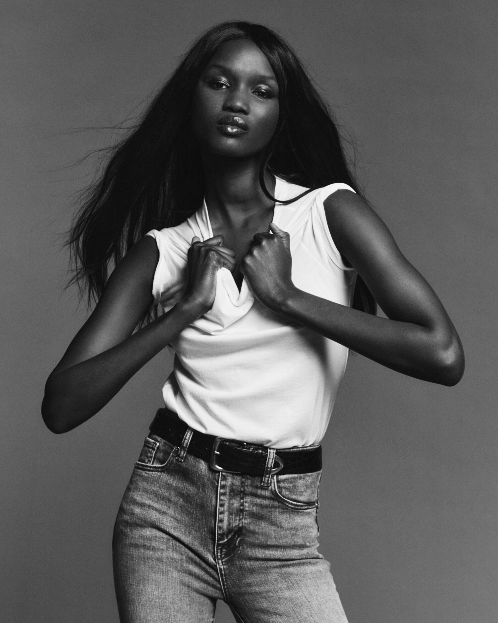 Black and white fashion portrait of a model with dark long hair wearing a white top and denim jeans against a grey background.