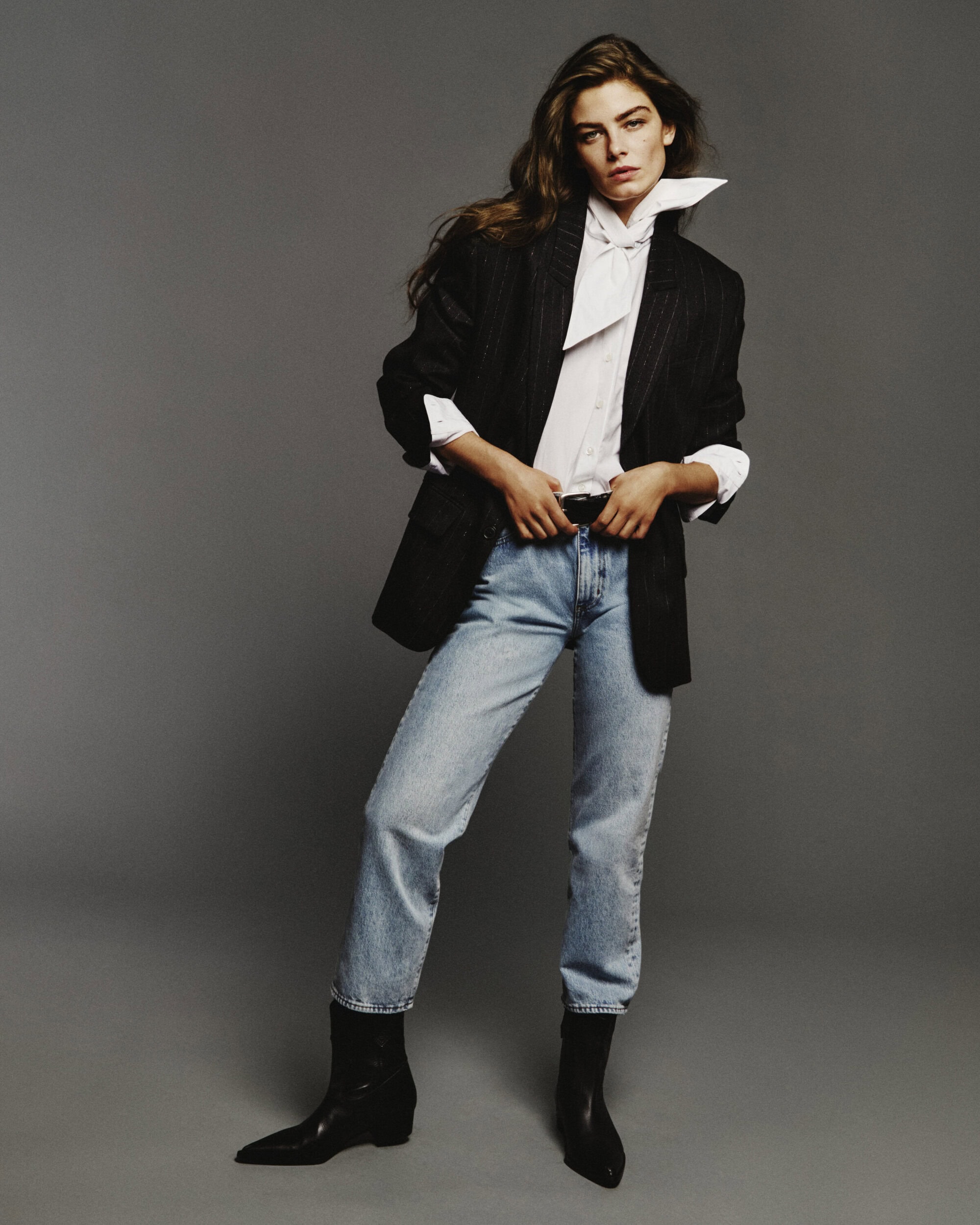 Model in a black blazer, white shirt, light wash jeans, and black boots against a grey studio backdrop.