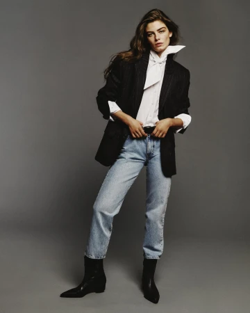 Model in a black blazer, white shirt, light wash jeans, and black boots against a grey studio backdrop.