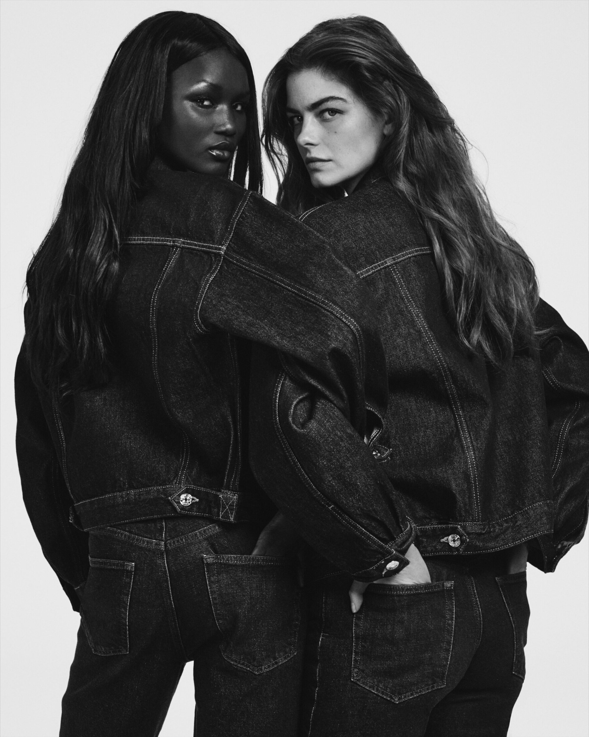 Two female models wearing matching dark denim jackets and jeans posing back to back against a white background.