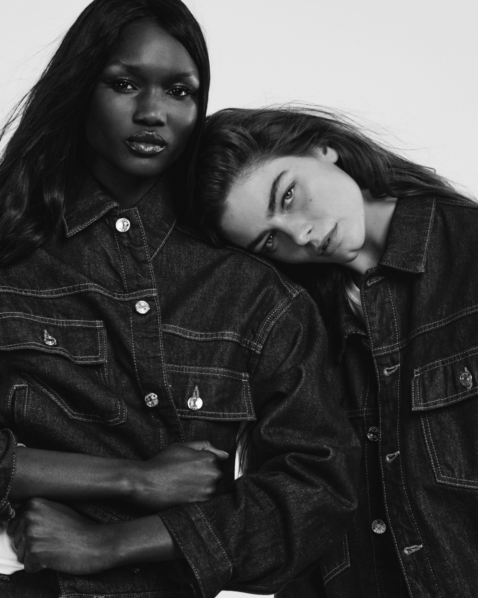 Two female models wearing dark denim jackets posing together against a plain light background in monochrome.