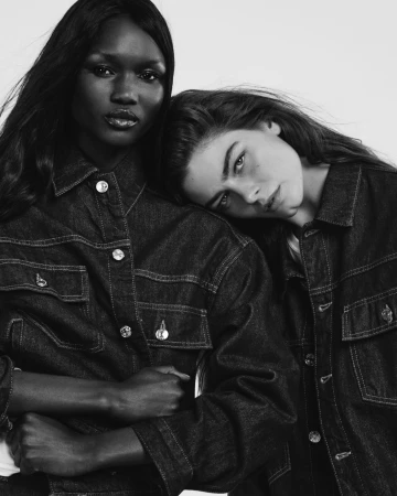 Two female models wearing dark denim jackets posing together against a plain light background in monochrome.