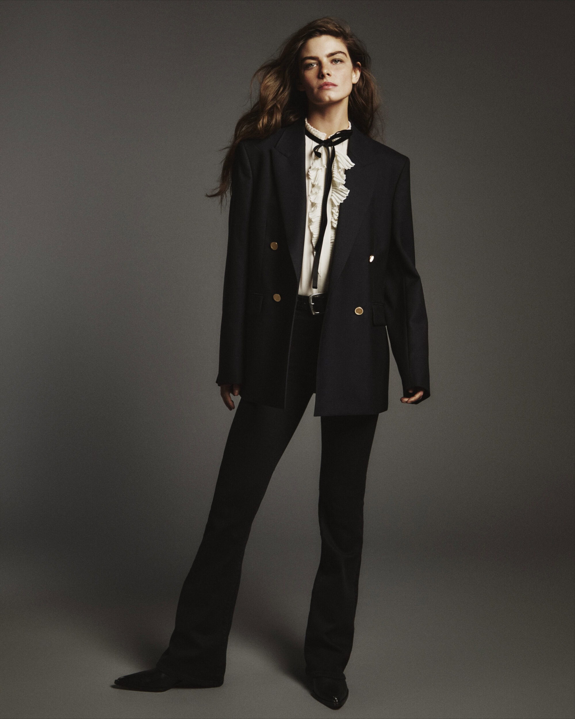 Model wearing a black blazer over a ruffled white blouse and black trousers against a neutral grey studio backdrop.
