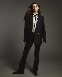 Model wearing a black blazer over a ruffled white blouse and black trousers against a neutral grey studio backdrop.