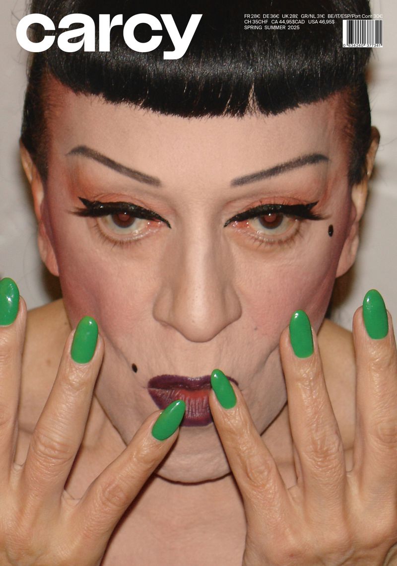 Close-up of a person with dramatic makeup and green nails, vintage-style haircut, and pale skin, against a neutral, soft-focus backdrop.