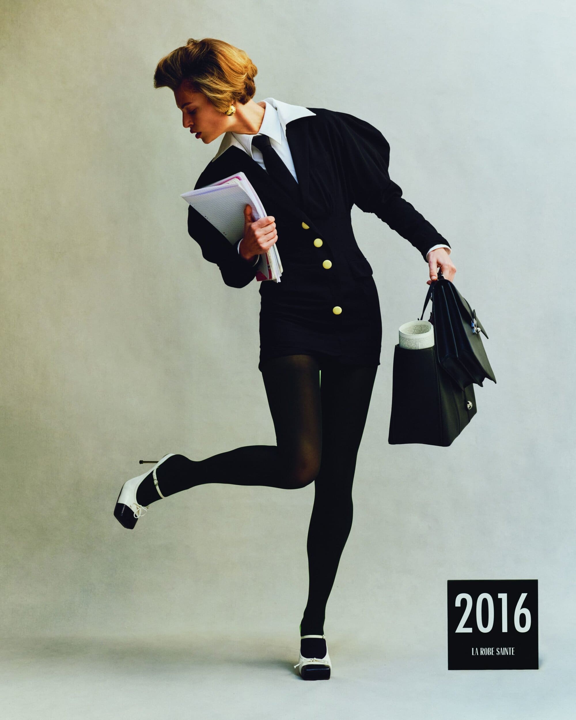 Woman in black jacket dress and white shirt holding folders and a bag against a plain background