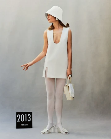 Model in white dress, bucket hat, tights and heels holding a bag against a neutral grey studio backdrop