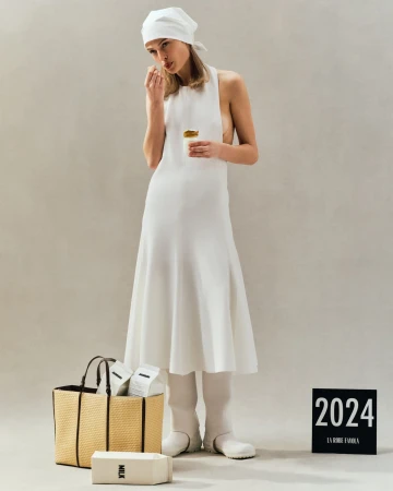 Model in white halter dress and headscarf holding a small jar, standing beside a woven tote bag on a neutral backdrop.
