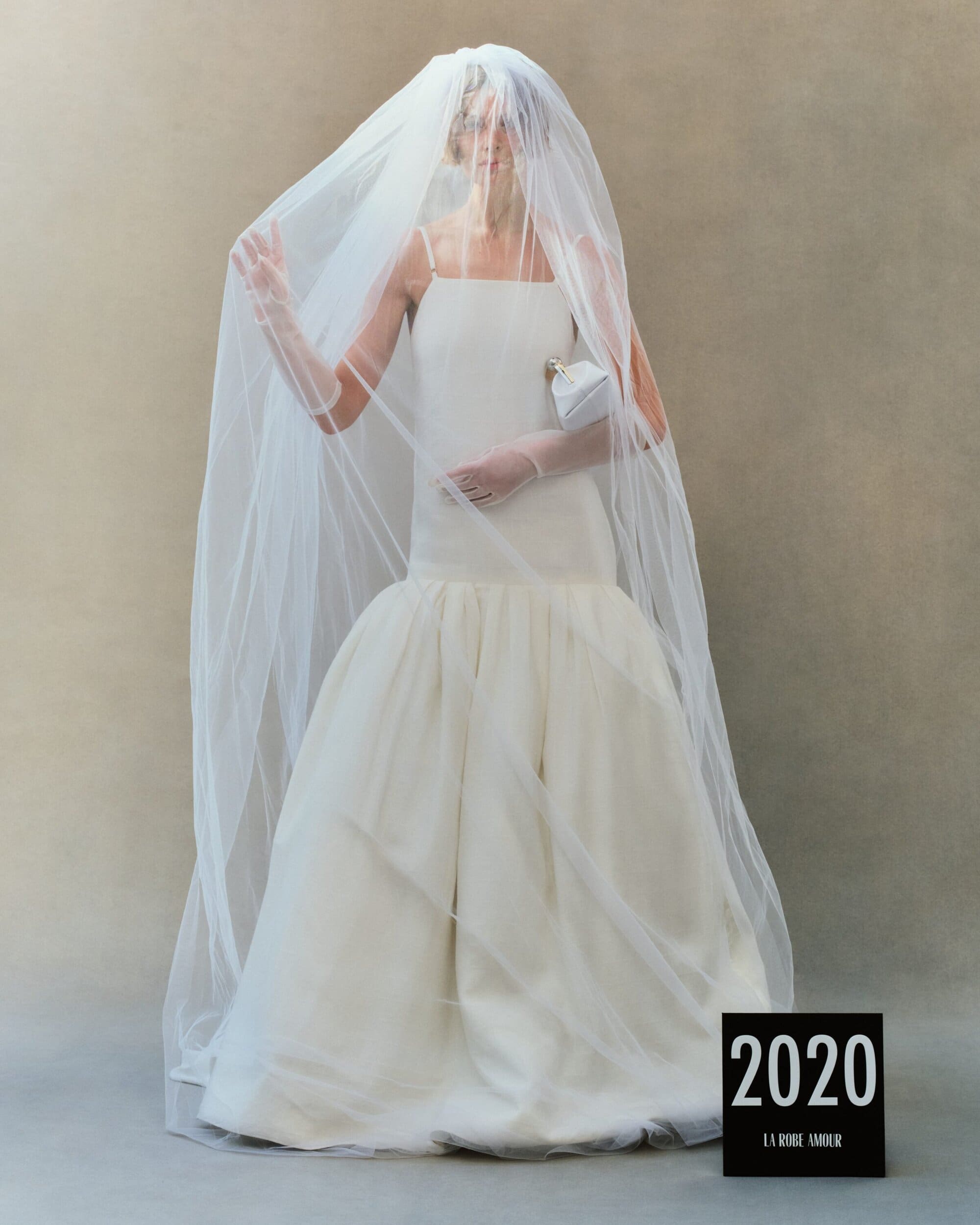 Model in white wedding dress wearing a large sheer veil and holding a small handbag against a neutral grey studio background.