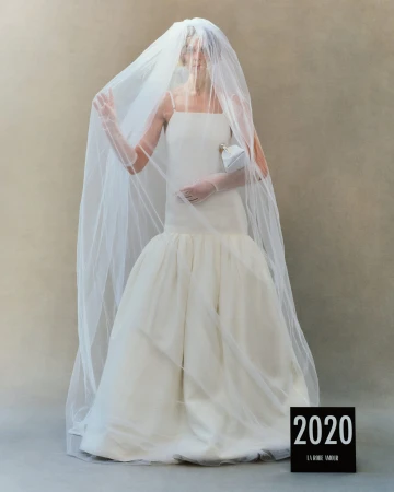 Model in white wedding dress wearing a large sheer veil and holding a small handbag against a neutral grey studio background.
