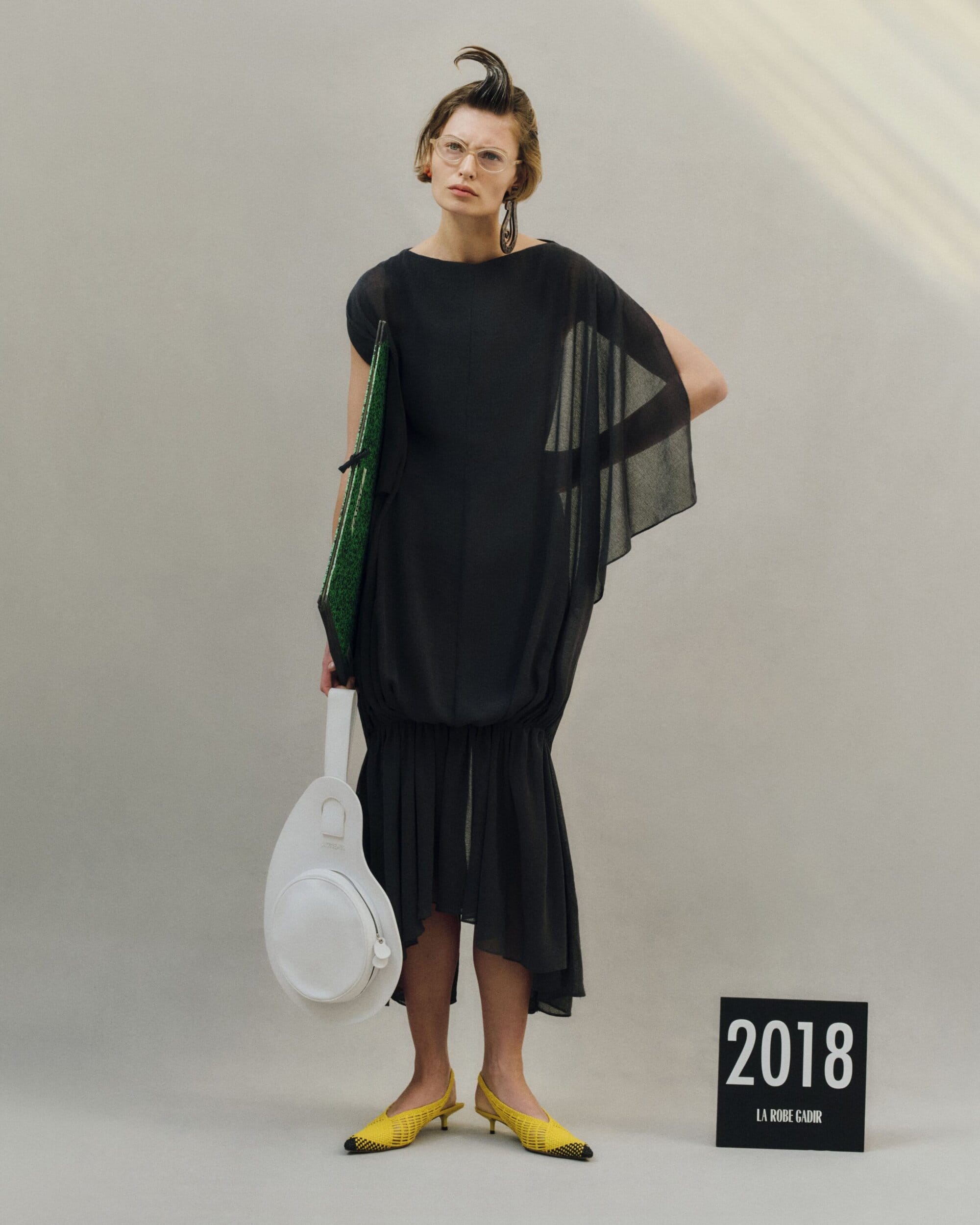 Model in a black dress holding a green folder and white handbag, wearing yellow shoes against a grey seamless background