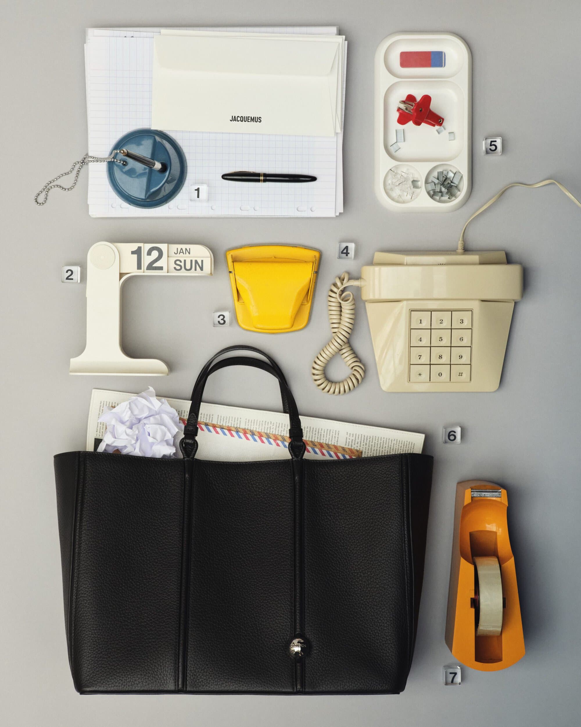 Overhead flat lay shot of office stationery, vintage telephone, and a large black leather handbag on grey backdrop