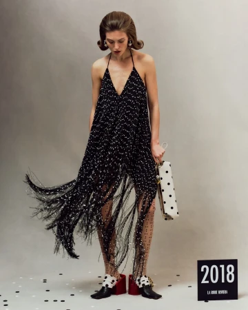 Model in a black fringe halter dress holding a white polka dot handbag against a neutral grey studio background